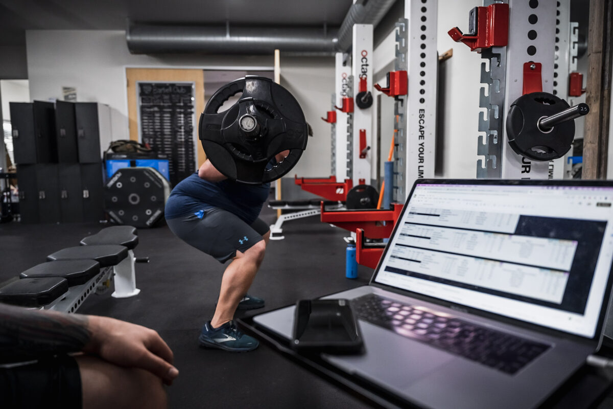 A client mid-squat at Training Space with a laptop and iPad in frame — the coach capturing training data live during the session.